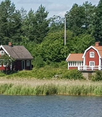 Lantlig scen med en slänt mellan två hus vid vattnet, omgivna av grönska.