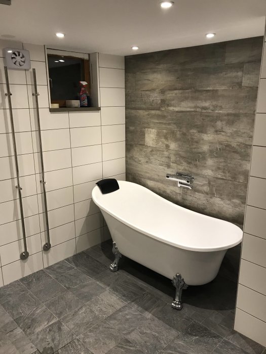 Nyinstallerat fristående badkar med retrostil ben i ett renoverat badrum med gråa kakelväggar och mörkgrått klinkergolv.