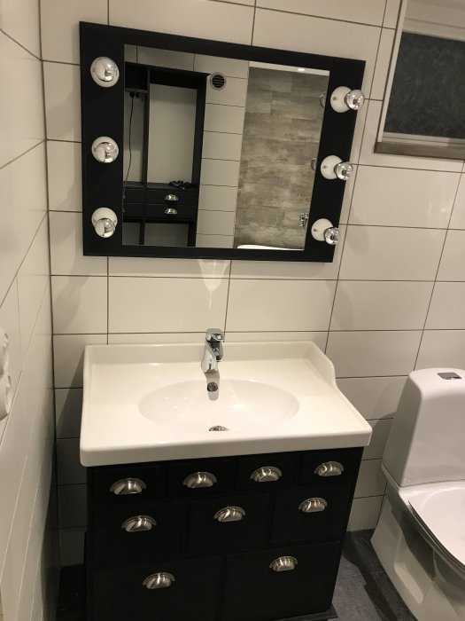 Badrum med svart handfatsskåp och handfat, spegel med omgivande lampor, och del av toalett.