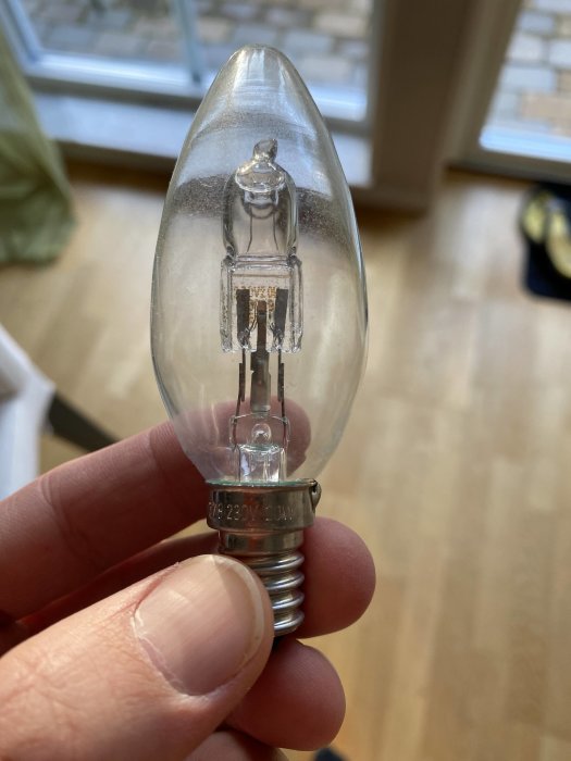 Hand håller en Osram Classic halogenlampa med tydlig märkning "230V 20W" framför ett fönster.