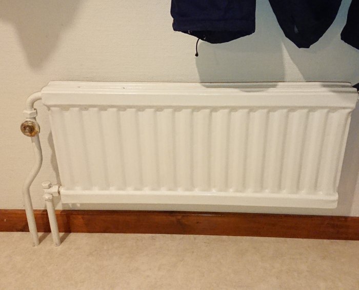 Vit radiator utan reglage mot en vit vägg med synlig röranslutning och parkettgolv.