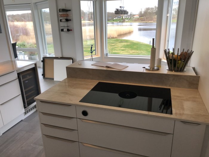 Kök under renovering med modern induktionshäll, ljusa skåp, marmorliknande bänkskivor och utsikt över sjö.