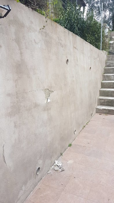 Skadad betongvägg med lossnat ytskikt och synligt glasfibernät vid sidan av en trappa.