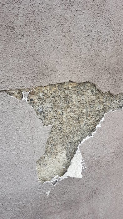 Skadad vägg där ytskiktet har släppt, exponerad betong och rester av glasfibernät ses.
