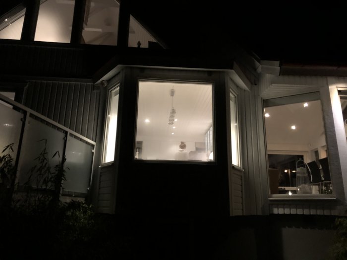 Kvällsbild av ett upplyst hörnfönster på hus där en person är synlig inuti.