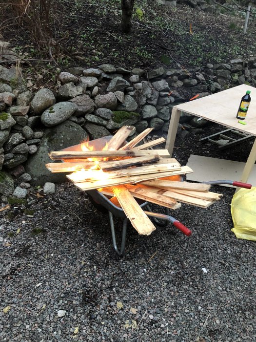 Eld upptänd på en skottkärra full med träplankor vid en stenmur, med ett bord och en flaska bredvid.