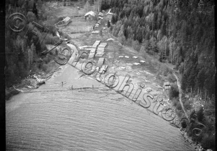 Svartvit flygbild från 1930-talet över en by vid en sjö med timmerstockar i vattnet.