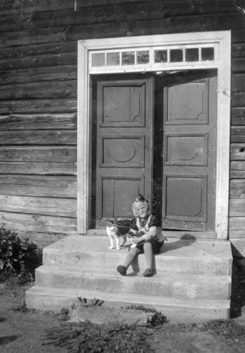 Gammalt svartvitt foto av ett barn som sitter med en katt framför en trädörr på 1930-talet.