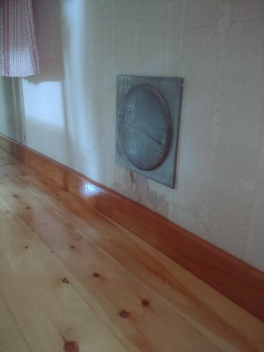 Ventil i en vägg nära golvet i ett rum med trägolv, isbildning synlig på ventilen.