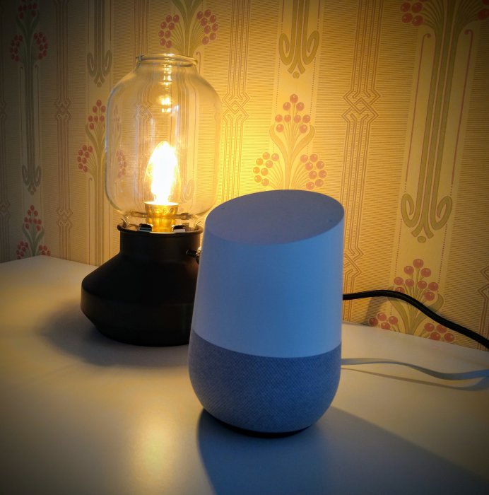 En smart högtalare från Google Home bredvid en tänd lampa på ett bord framför retro tapet.