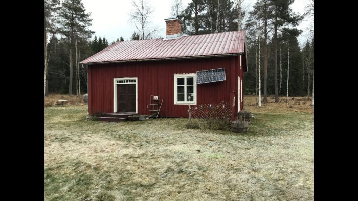 Rödmålat litet timmerhus med vita knutar och fönster, omgivet av frostigt gräs och skog i bakgrunden.