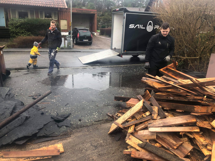 Två personer och ett barn som flyttar hopkapade trädelar framför en släpvagn och en söndertagen svart asfaltyta.