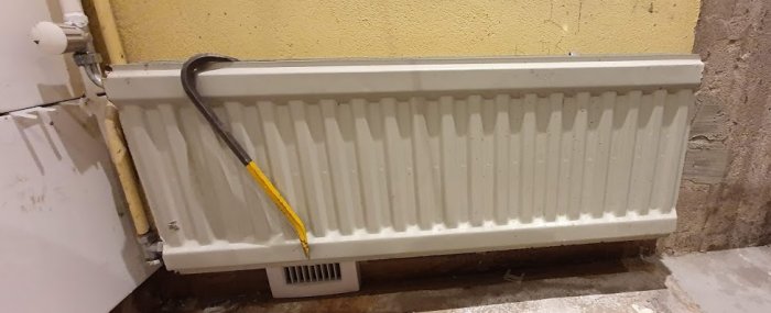 Ett läckande vitvattenradiator element intill en gul vägg med synliga rör och en värmefördelningsplåt undertill.