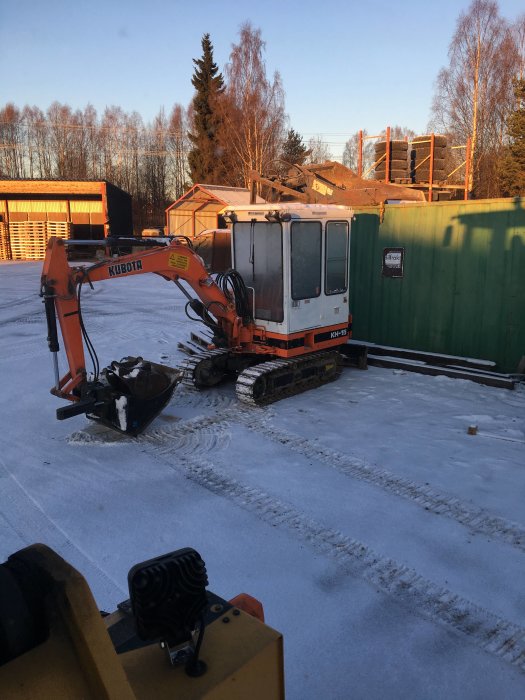 Orange Kubota grävmaskin på snöig mark bredvid gröna containrar och byggställning i bakgrunden.