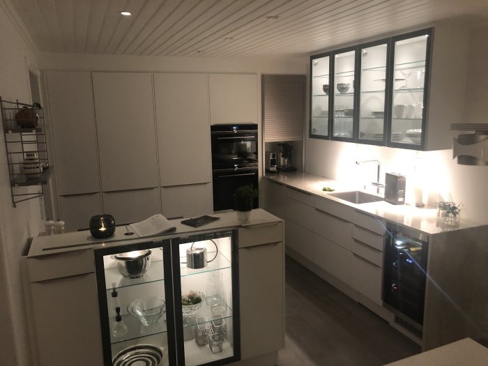 Modernt kök med vita skåp, inbyggda vitvaror och belysning under överskåpen.