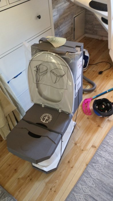 Ny toalettstol förberedd för montering på ett trägolv nära badrumsskåp och verktyg.