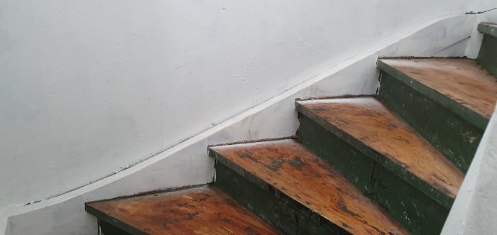 Trappa med sliten grön färg och trädetaljer med en springa mellan sidstycke och vit vägg.