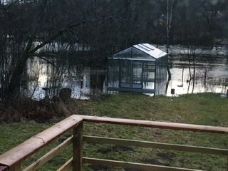 Översvämmad trädgård med vatten kring ett växthus, sett från en träaltan.