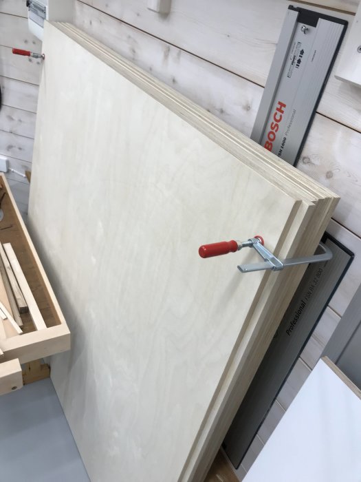 Flera skivor av finplywood staplade på varandra i en verkstad med en G-klämma och en Bosch-sågguide i bakgrunden.