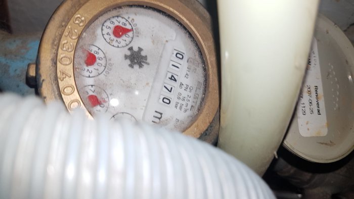 Närbild av en gammal vattenmätare med smutsig display intill vita rör och slangen.