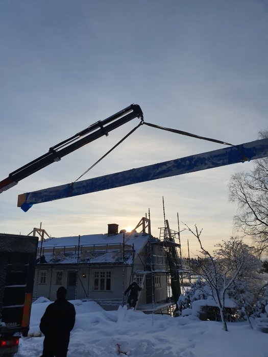 Kran som lyfter en balk på en byggarbetsplats under vintern med snö och en solnedgång i bakgrunden.