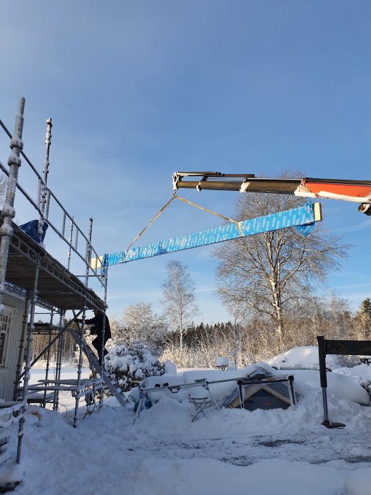 En stor blå balk som lyfts på plats av en kran vid ett byggnadsställning i vinterscen.