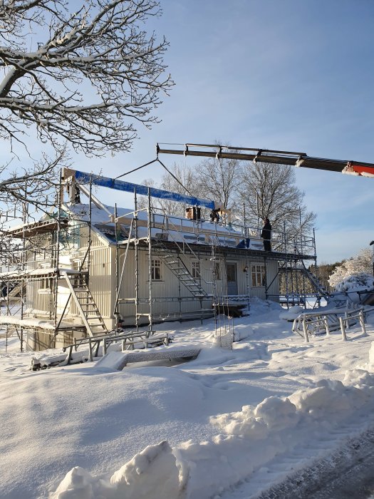 En balk lyfts på plats till ett hus under konstruktion i ett snötäckt landskap.