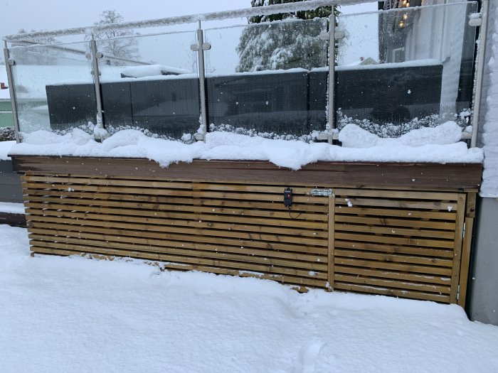 Snötäckt trädgårdsdäck med en delvis synlig glasräcke och träavskärmning, i ett snöigt landskap.
