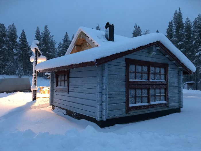 Trästuga täckt med snö vid skymning, omgiven av snötäckt landskap och snöklädda träd.