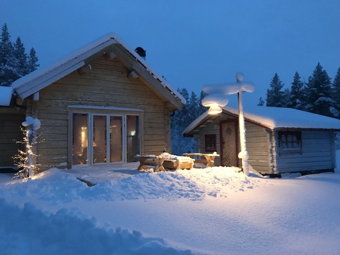 Trähus och förråd täckta med snö i skymningen med utomhusbelysning och omgiven av snötäckt landskap.