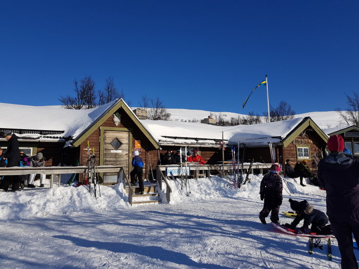 Människor njuter av solskenet utanför en snötäckt stuga med skidor, på en klar vinterdag.