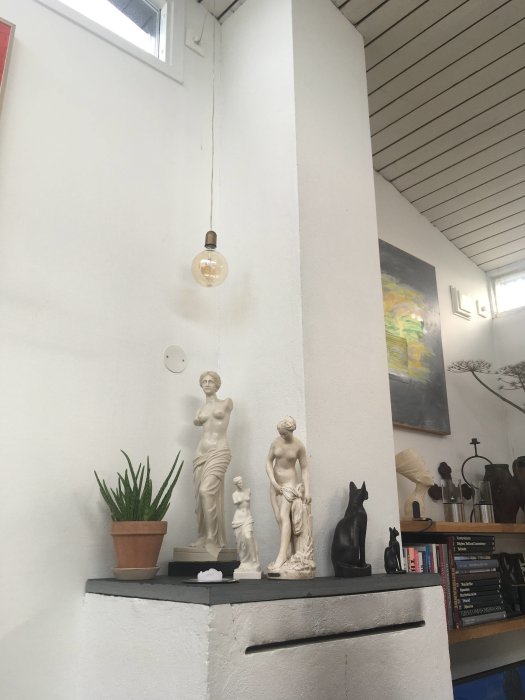 Statuer og planter på en hylle med hvit vegg, hengende lyspære, og ei bokhylle med bøker i bakgrunnen.