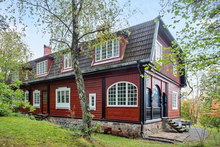 Välbevarat traditionellt svenskt rött hus med vita knutar och svart plåttak omgivet av grönska.