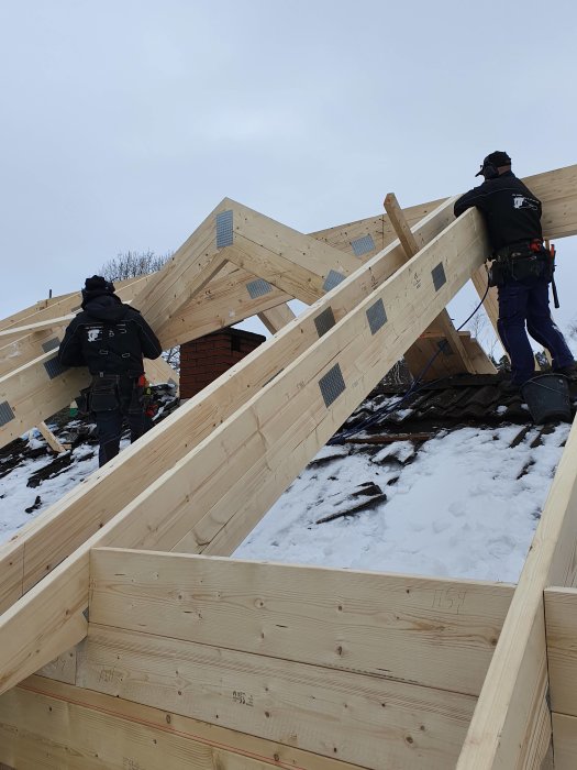 Två arbetare monterar trästommen till ett hus under konstruktion på en snötäckt byggplats.