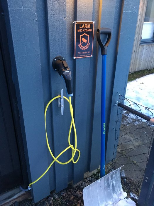 Hemmagjord laddstation för elbil fäst med buntband på en blå yttervägg, bredvid en skylt och snöskyffel.