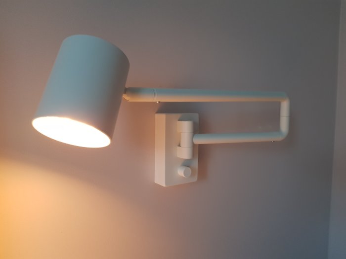 Modernt väggmonterat ljus med tänd lampa och justerbar arm mot en grå vägg.