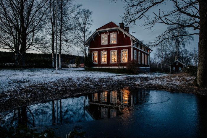 Rött hus med tända fönster och spegling i en frusen vattensamling vid skymning.