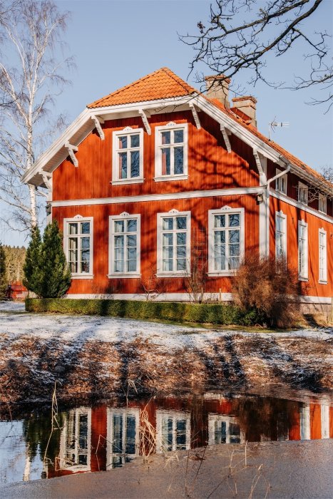 En röd timmerbyggnad med snedtak och vita fönster speglar sig i en vattenpöl omgiven av gräs med snöfläckar.