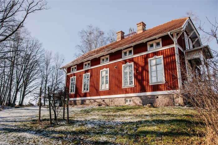 Rödmålad trävilla med vita fönster och tegeltak i lantlig miljö med lätt snötäcke på marken.