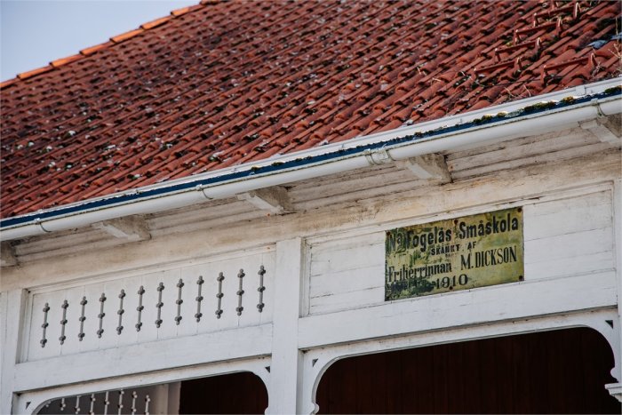 Del av gammal byggnad med vit träfasad, snidade balkongdetaljer och skylt som säger 'Nätögla Smäskola 1910' under ett tegeltak.