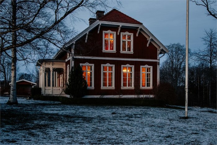 Rödmålat trähus med tända fönster och snöfläckig gräsmatta i skymningen.