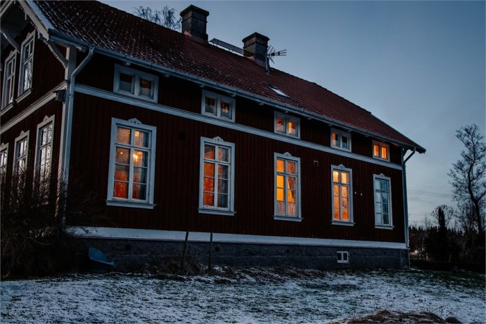 Skymningsvy av ett rött trähus med tända fönster som speglar en varm atmosfär mot en snöströdd trädgård.