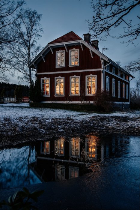 Röd traditionell svensk villa med tända fönster och spegling i en vattenpöl under skymningen.