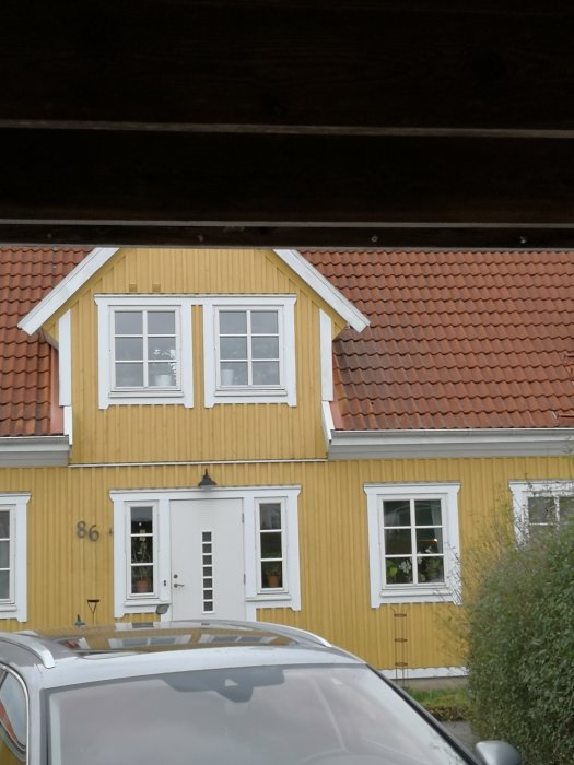 Gult hus med spröjsade fönster, ett med horisontella spröjs, under en träbjälke.