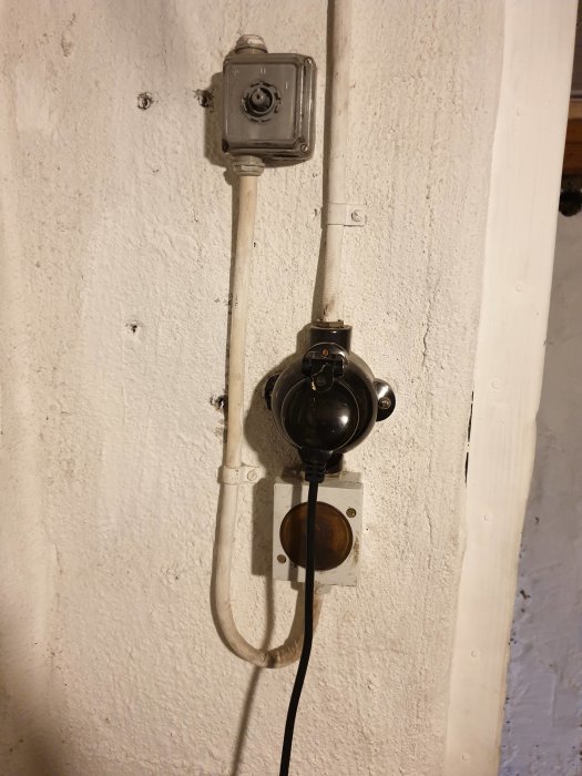 Gammal elektrisk installation med kabel och svart rund enhet på vit vägg, eventuellt kopplad till äldre värmepanna.