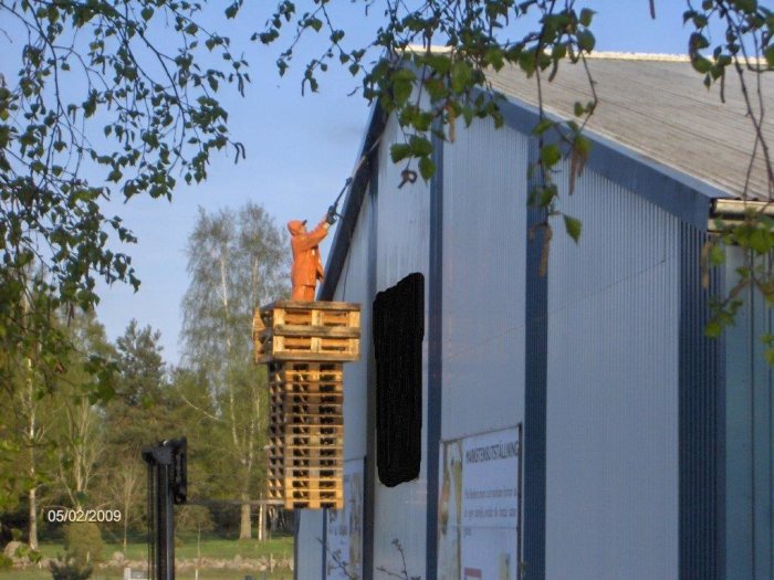 Person i orange skyddsdräkt på en pallkrage staplad på en gaffeltruck bredvid en industribyggnad.