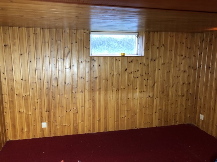 Gillestuga med furupanel på väggarna och röd heltäckningsmatta på golvet före renovering.