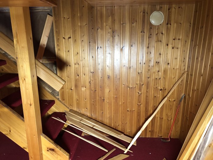 Träpaneler och röd heltäckningsmatta i ett rum under renovering, med borttagen panel och synliga träreglar.