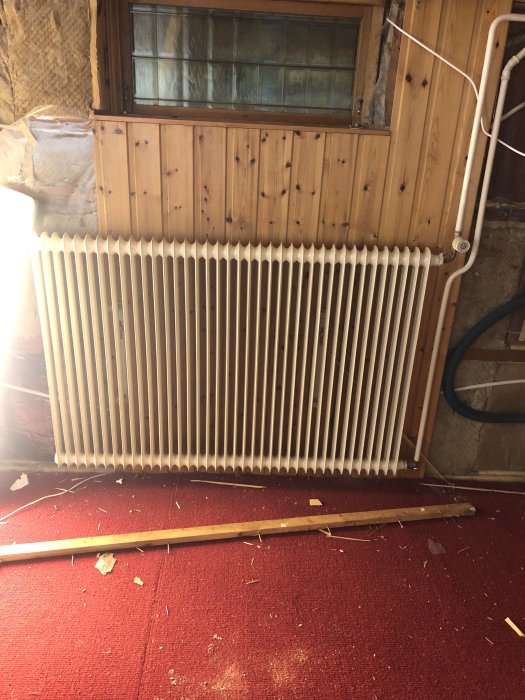 Renoveringsbild av gillestuga med furuvägg och element framför ett fönster, över heltäckningsmatta och spillvirke på golvet.