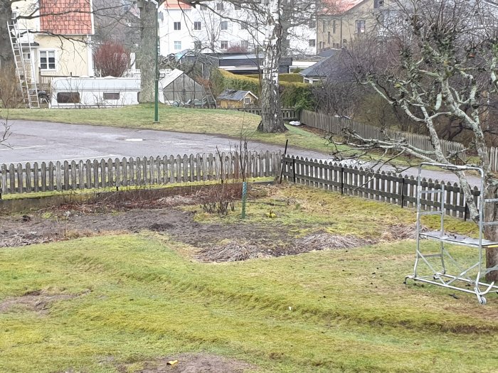 Vy över ett trasigt staket längs en gräsplätt med parkeringsområde och träd i bakgrunden.